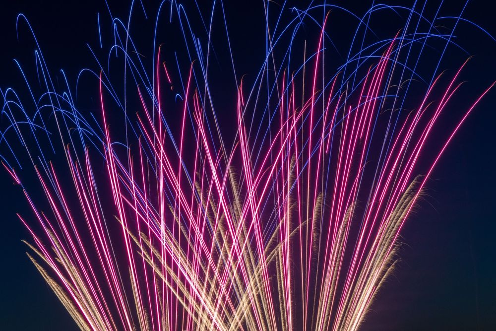 Thin streaks of light formed by fireworks in long-exposure photography. They're shooting up from the ground and curving downward into the night sky. They're yellow at the base, pink in the middle, and blue on top.