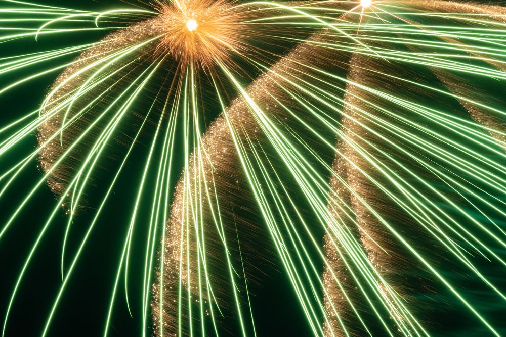 Long-exposure firework photo. A yellow ball bursts into yellow sparkles and green streaks of light which look like lasers.