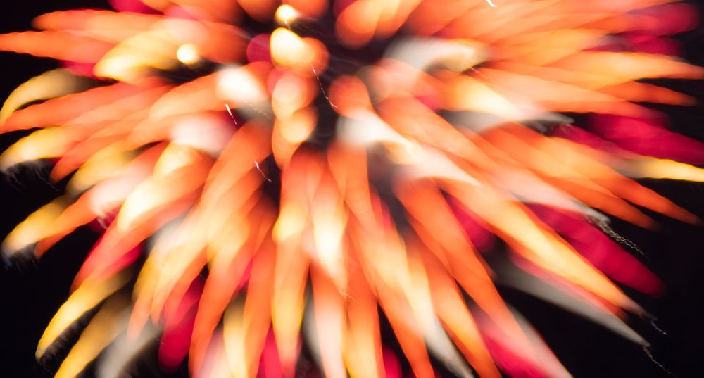 Long-exposure defocused and refocused picture of orange and red firework sparkles. The image is soft and looks like a bright orange and red flower with spiky pedals.