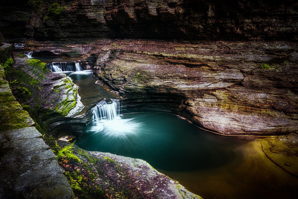 Photo taken of a couple small waterfalls emptying into a larger natural pool at the bottom of the cavern type area. 