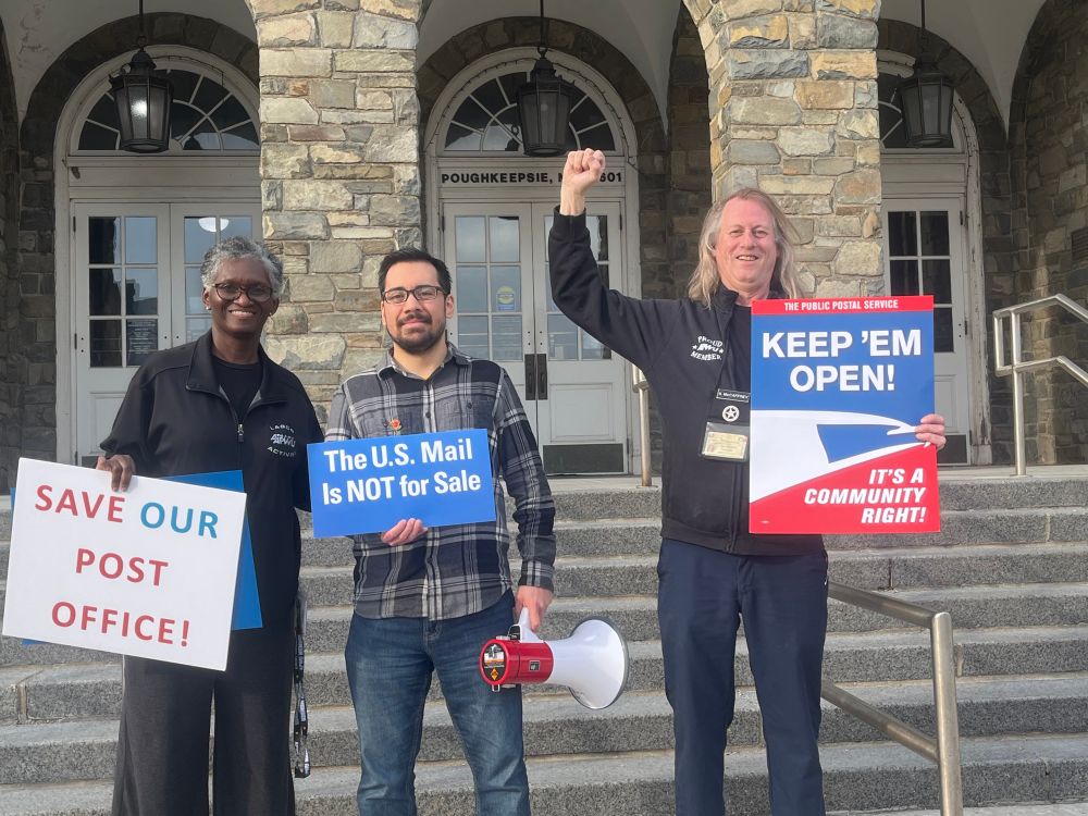 I’m rallying with APWU members outside the Poughkeepsie Post Office. I’m holding a megaphone and a sign that says “The U.S. Mail is NOT for Sale”.