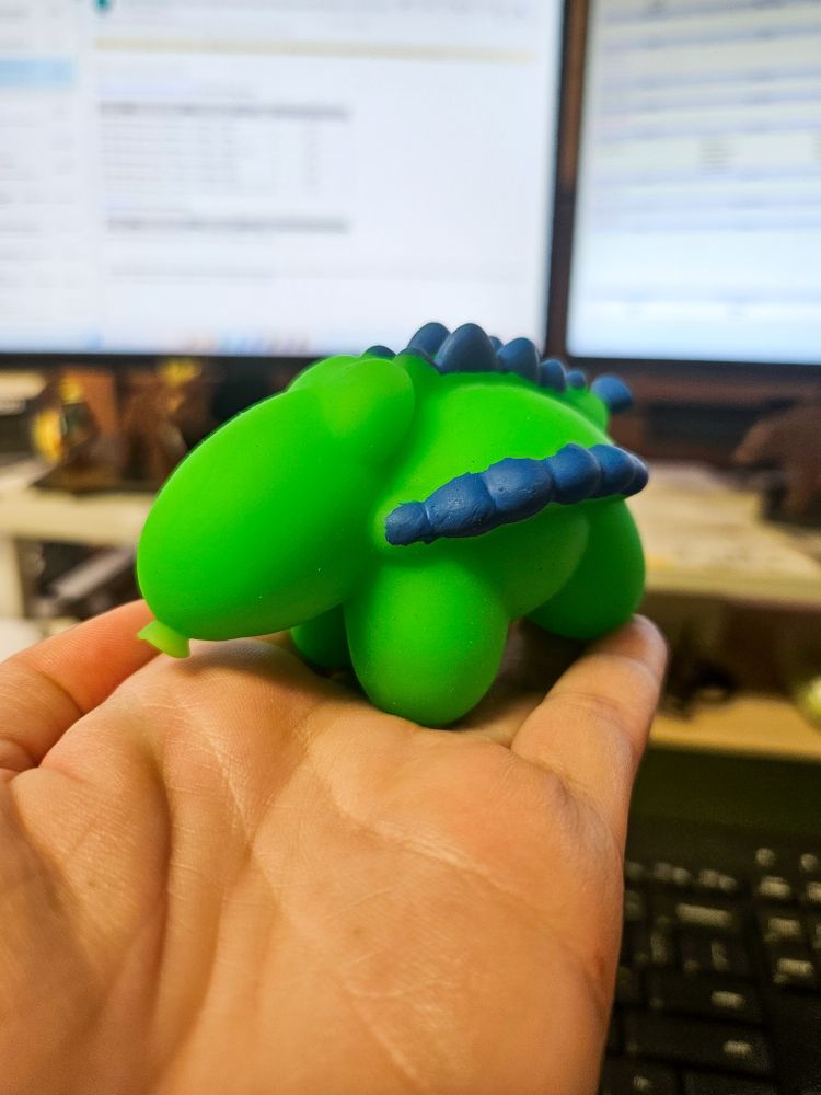 A green and blue toy that looks vaguely like an ankylosaurus as a balloon animal. The background is a work desk with a keyboard and two screens.