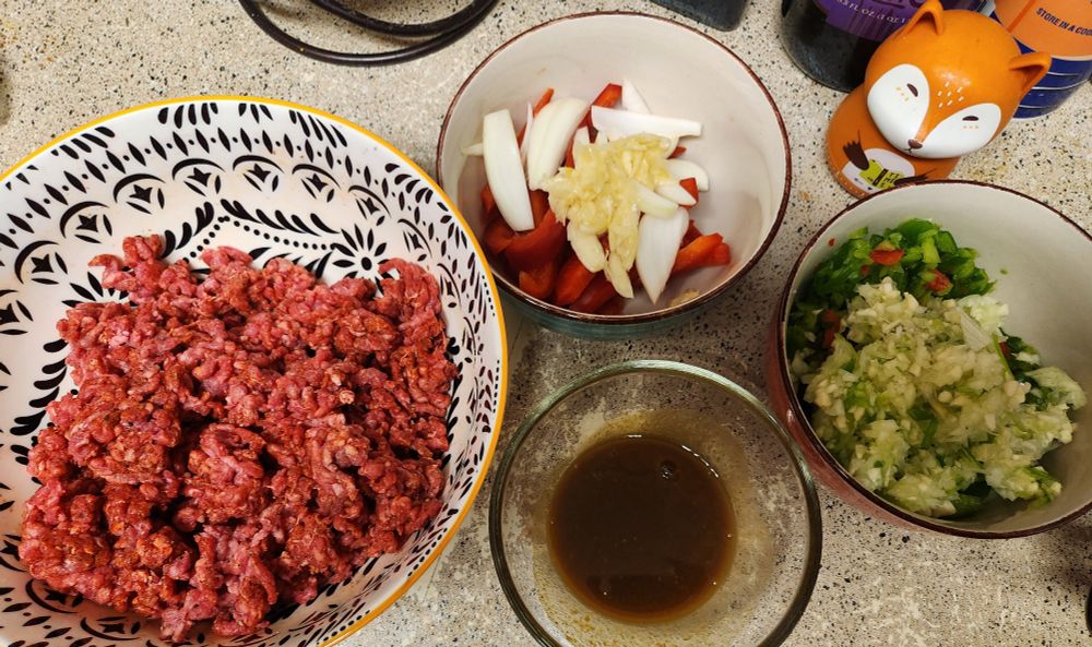 Prep for homemade sloppy joes. Bowls of seasoned ground beef, a worchestershire sauce blend, and finely chopped onions and bell peppers. A fourth bowl has chunks of onion, red bell pepper, and crushed garlic to season sautéed cabbage.