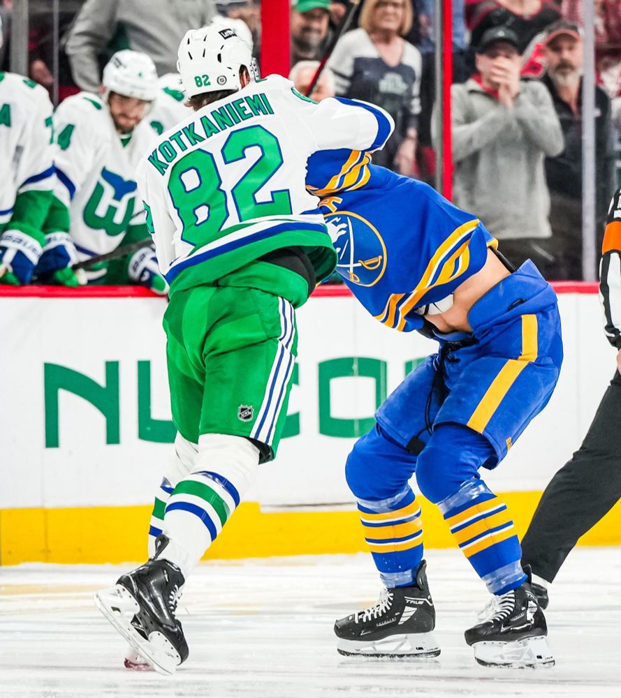One hockey player in a white jersey and green pants fighting another player in all blue 