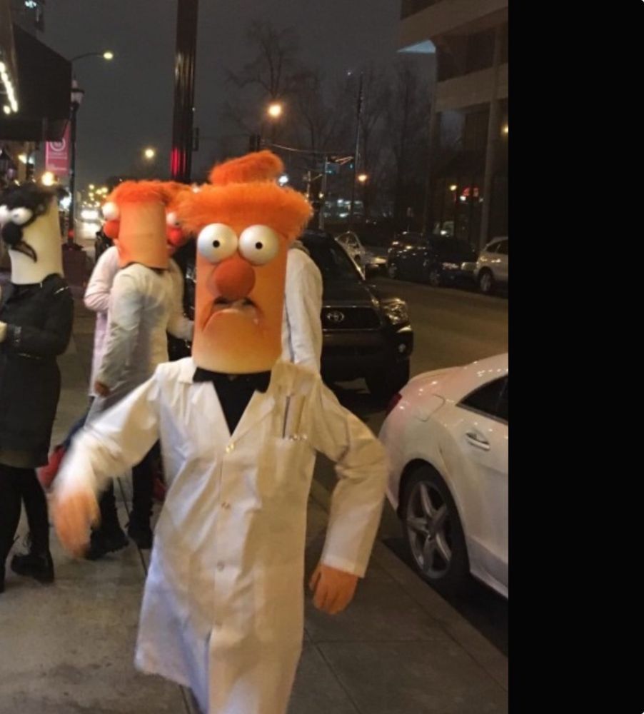 Pack of Beakers at night. Alt alt text: person in wearing Beaker head and lab coat striding toward camera with people in Beaker costumes on a city street at night in background