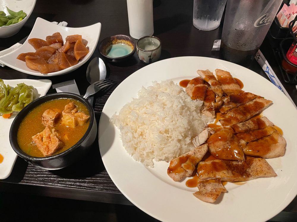 A dinner setting at a Japanese restaurant. The main plate is white rice with grilled chicken and a brown sauce. Next to it is a bowl of brown soup with fried tofu. Also in the picture are some greens, edamame, pickled radish, and a tokkuri of sake. 