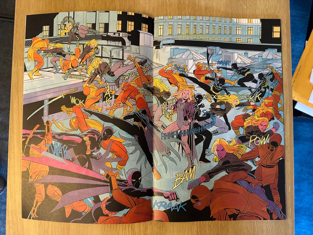 Photo of an open comic book on a table. Two-page spread from Birds of Prey (2023) issue 1. An action scene unfolds between Black Canary, Cassandra Cain and a group of identically costumed assailants. The movement of the fight is shown through repetition of the primary figures, with Dinah moving top left to bottom right as Cass move top right to bottom left. Both meet just right of center, back to back, and take S-curves towards center bottom where they issue matching patches in rough symmetry towards bottom center. Colors are simplified in a way that makes each set of figures easily trackable, gives the opponents a unified look and makes the background, of a rooftop and surrounding buildings, maintain detail while staying out of the way so the figures pop out clearly. Cass’ sound effects are in blue, Dinah’s in yellow—brighter variations of the top colors used for the BG and matching highlights from each figure. Also there is much cool-looking punching and kicking. 