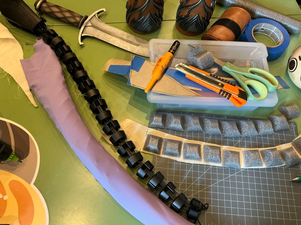 A table strewn with craft supplies, such as scissors, knives, pens, tape and a cutting mat. On the mat are some small pieces of foam laid out on a strip of fabric. To the side is the internal structure of a mechanical tail, and a ribbed purple sleeve, suggestive of a tiefling tail.