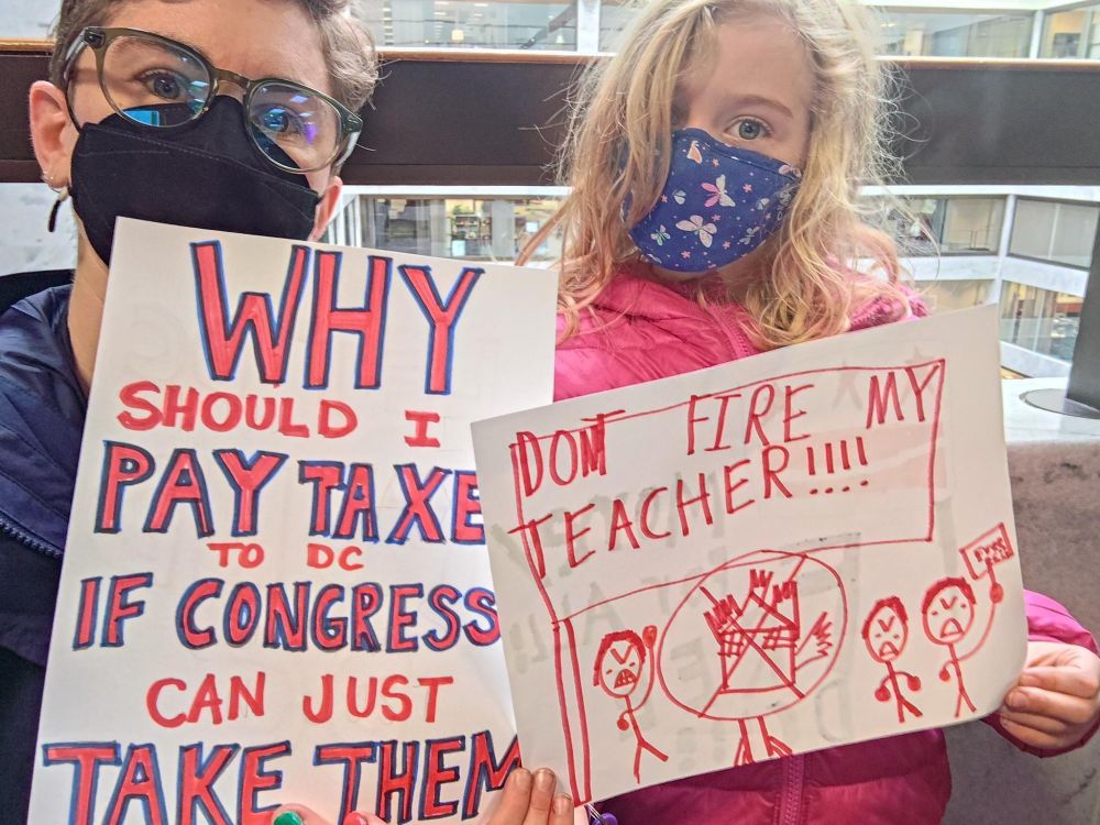 BHES mom in glasses and mask holds a sign that reads "Why should I pay taxes in DC if Congress can just take them?" and BHES scholar with mask and pink coat holds a sign that reads "DON'T FIRE MY TEACHER!!!!" in kid hand-writing with angry stick figures below.