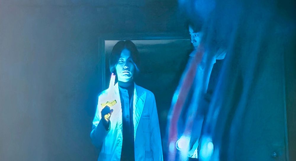 An Eun-young rests a light-up, plastic, toy sword on her shoulder as she looks towards a jelly in a dark-lit hallway. Hong In-pyo looks at her curiously, wondering what she is doing. They stand inside a doorway that leads to the hallway. This is from the kdrama School Nurse Files.