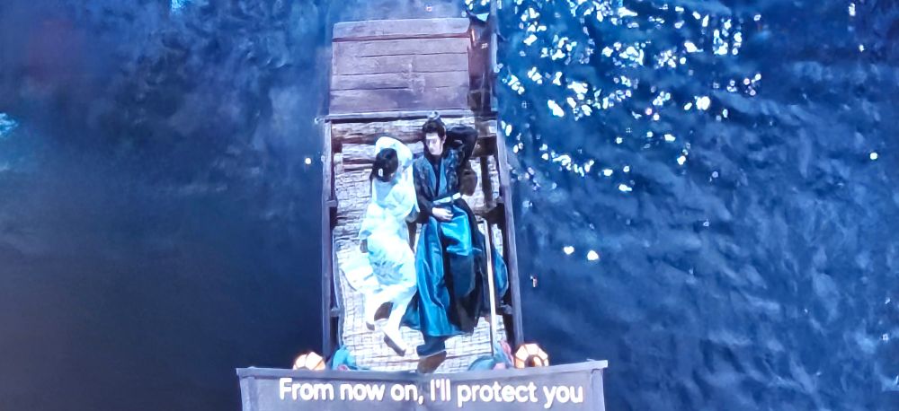 Fu YiXiao leans next to Feng Suige, who reclines on a boat. The boat is floating on deep blue water. They look relaxed and comfortable. Text says, "From now on I'll protect you." This is from the cdrama Fated Hearts.