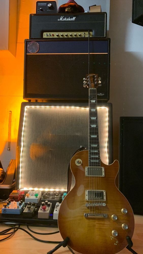 A Gibson Les Paul electric guitar in front of a Marshall 1987X amplifier with two cabinets and a pedalboard.