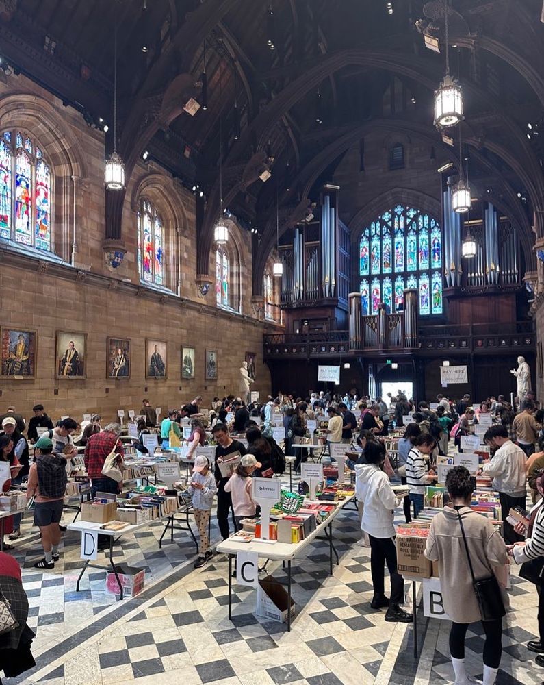 A University hall with tall sandstone walls, dark wood roofing, chequered tiles, stained glass windows, a carillon, and portraits of old white men on the walls. It’s filled with trestle tables covered with books and dozens of people.