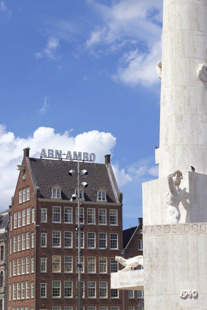 A shot of the National Monument in Amsterdam, with a tall traditional brick building in the background, set against a bright blue sky.