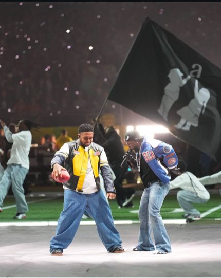 Kendrick Lamar and Dj mustard performing at the Super Bowl halftime show while dancers in the background hold a flag of two kids reaching for a lowercase letter “a”