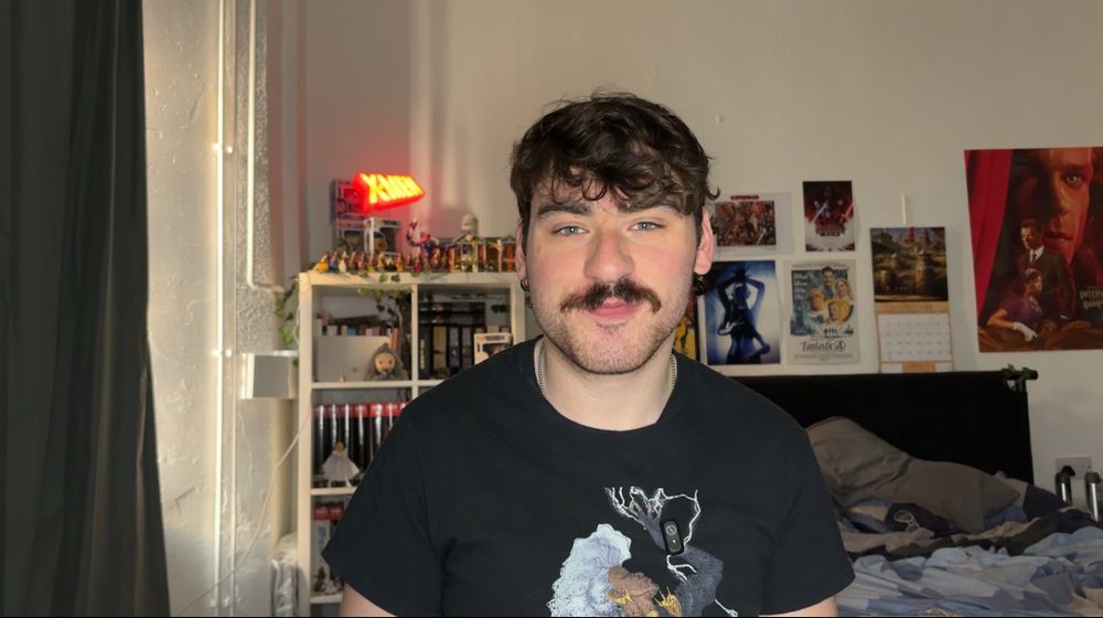 a man with a moustache and brown hair wearing a storm (Marvel hero) shirt. on the wall is a beyoncé poster, an interview with a vampire poster, an x-men poster etc.
