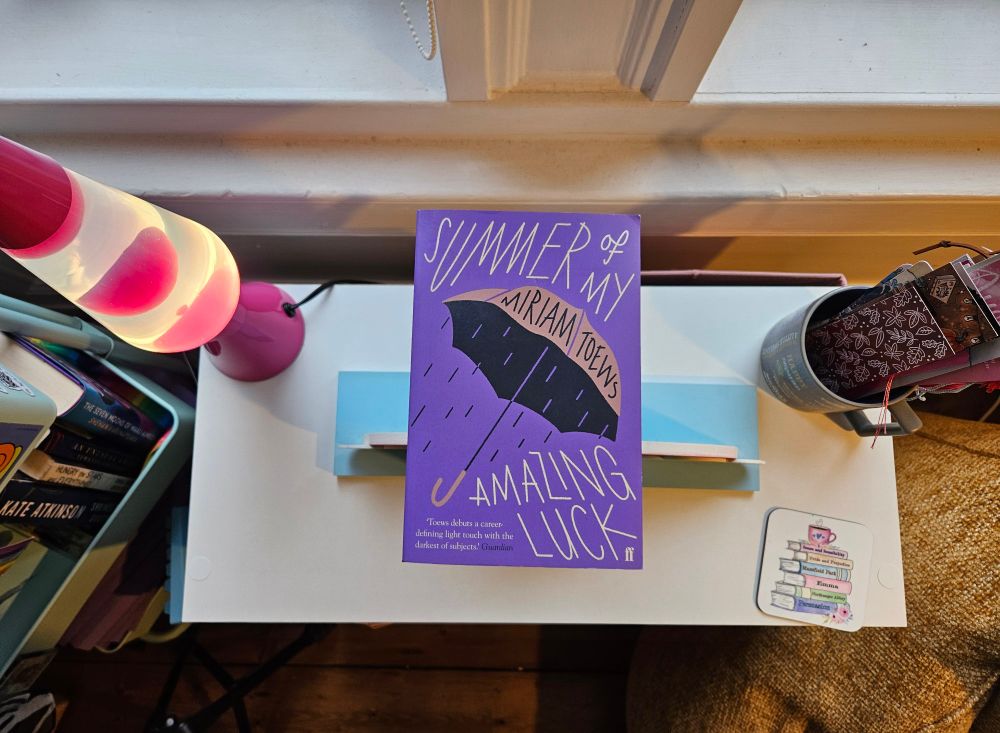 Picture of The Summer of My Amazing Luck by Miriam Toews on top of a small white bookcase surrounded by a pink lava lamp, a mug full of bookmarks, and a white coaster depicting a stack of Jane Austen novels.