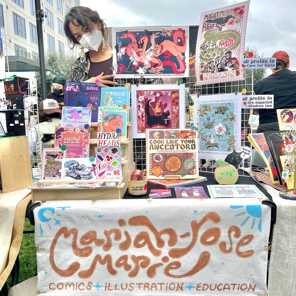 a masked brown human with smiling eyes stands next to a glorious table spread of zines, prints and books.
