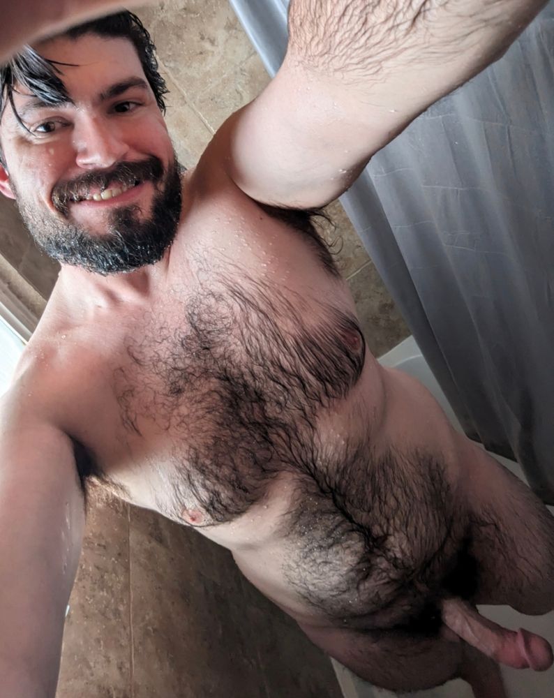 Dingo standing naked in the shower with wet hair and skin. Holding the camera at a down facing cocked angle and giving a cheeky smirk