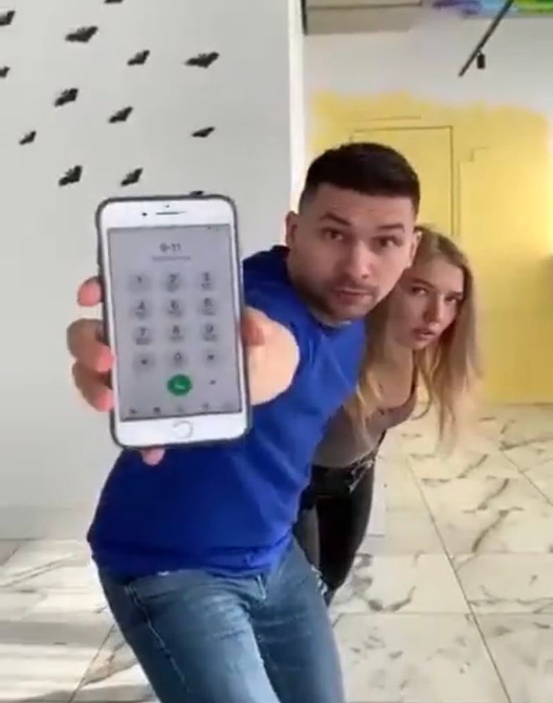 Man guarding a woman behind him while holding out his phone, the screen showing that he dialed 9-1-1