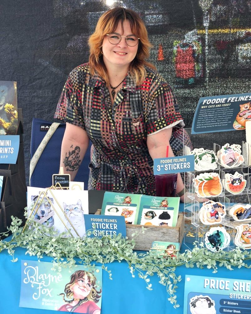 Myself, a plus sized woman in a colorful jumpsuit behind a booth offering cat stickers, mini prints and several labels advertising myself. 