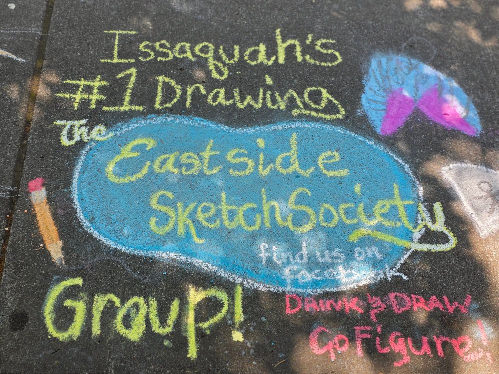 A chalk art drawing of the Eastside Sketch Society logo which has yellow font and a blue bean shaped background. It quotes Drink & Draw, Go Figure and that we are the #1 drawing group in Issaquah! 