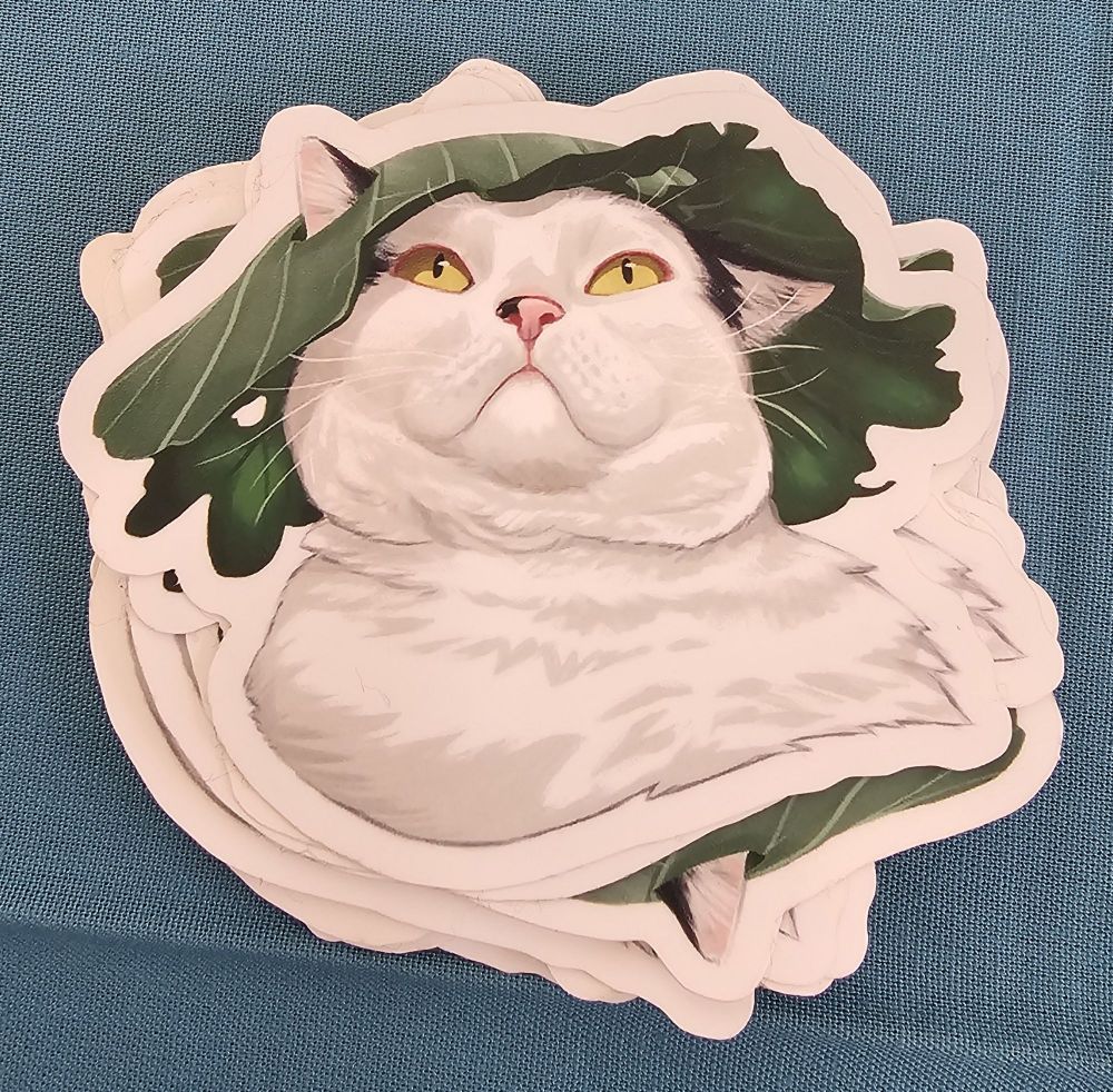 A cat sticker featuring a round white and black cat with yellow eyes and a Cabbage leaf on his head. 