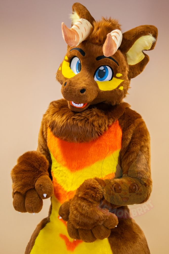 A full-body picture of a dragon fursuit. The dragon is primarily brown with yellow and orange markings, and front facing horns that are beige with more beige-pink accent markings near the tips of the horns. They have a cute, happy expression as they raise their paws in front of their belly.