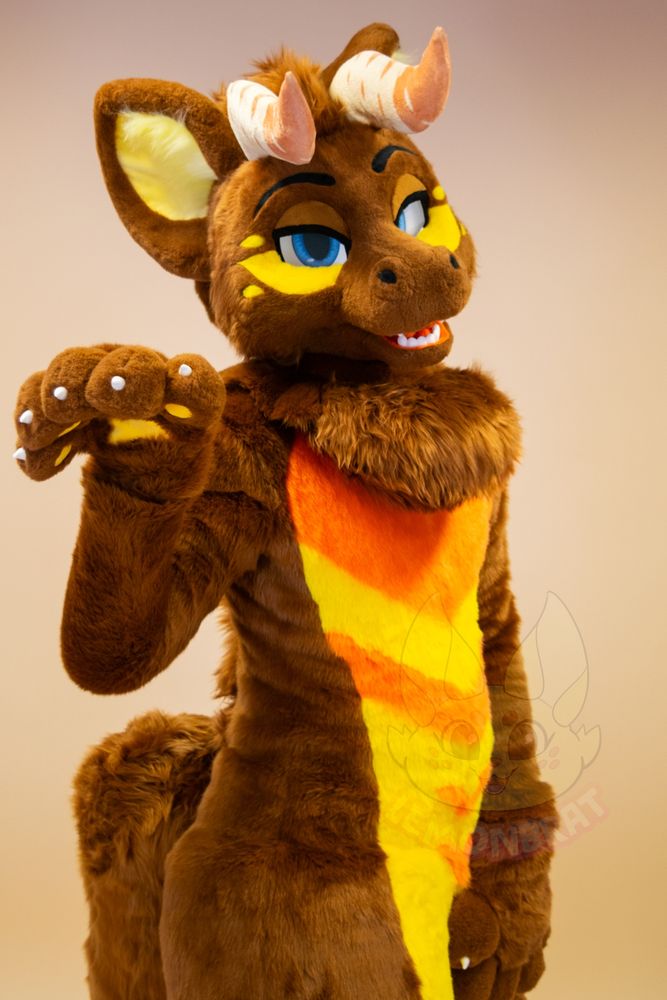 A half-body picture of a dragon fursuit. The dragon is primarily brown with yellow and orange markings, and front facing horns that are beige with more beige-pink accent markings near the tips of the horns. They give a sassy kinda look with half-lidded eyes. One paw is raised in front of them, almost mockingly limp-wristed, while they slightly tilt their head to the side.