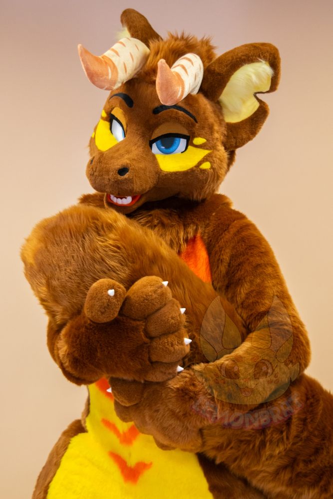 A half-body picture of a dragon fursuit. The dragon is primarily brown with yellow and orange markings, and front facing horns that are beige with more beige-pink accent markings near the tips of the horns. They give a sassy kinda look with half-lidded eyes while they cuddle their long, cat-like tail in both paws against their chest.