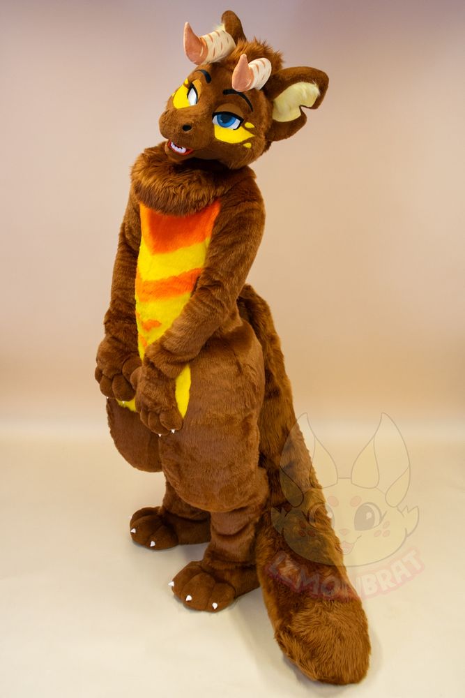 A full-body picture of a dragon fursuit. The dragon is primarily brown with yellow and orange markings, and front facing horns that are beige with more beige-pink accent markings near the tips of the horns. They have a long, cat-like tail that lies along the floor behind them, and give a sassy kinda look with half-lidded eyes. They have their hand paws on their waist with their head tilted to the side.