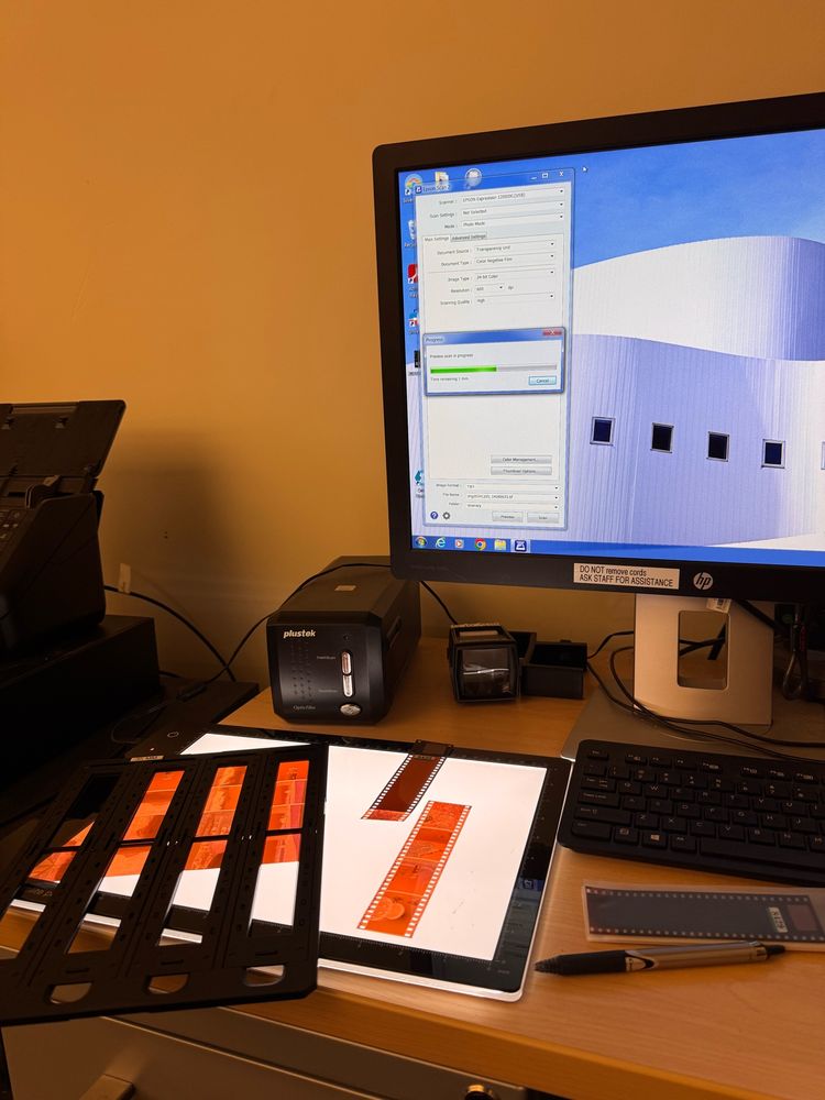 A light box sits on a desk in front of a computer screen with mounted 35mm negatives on it. The computer shows that it is scanning files.