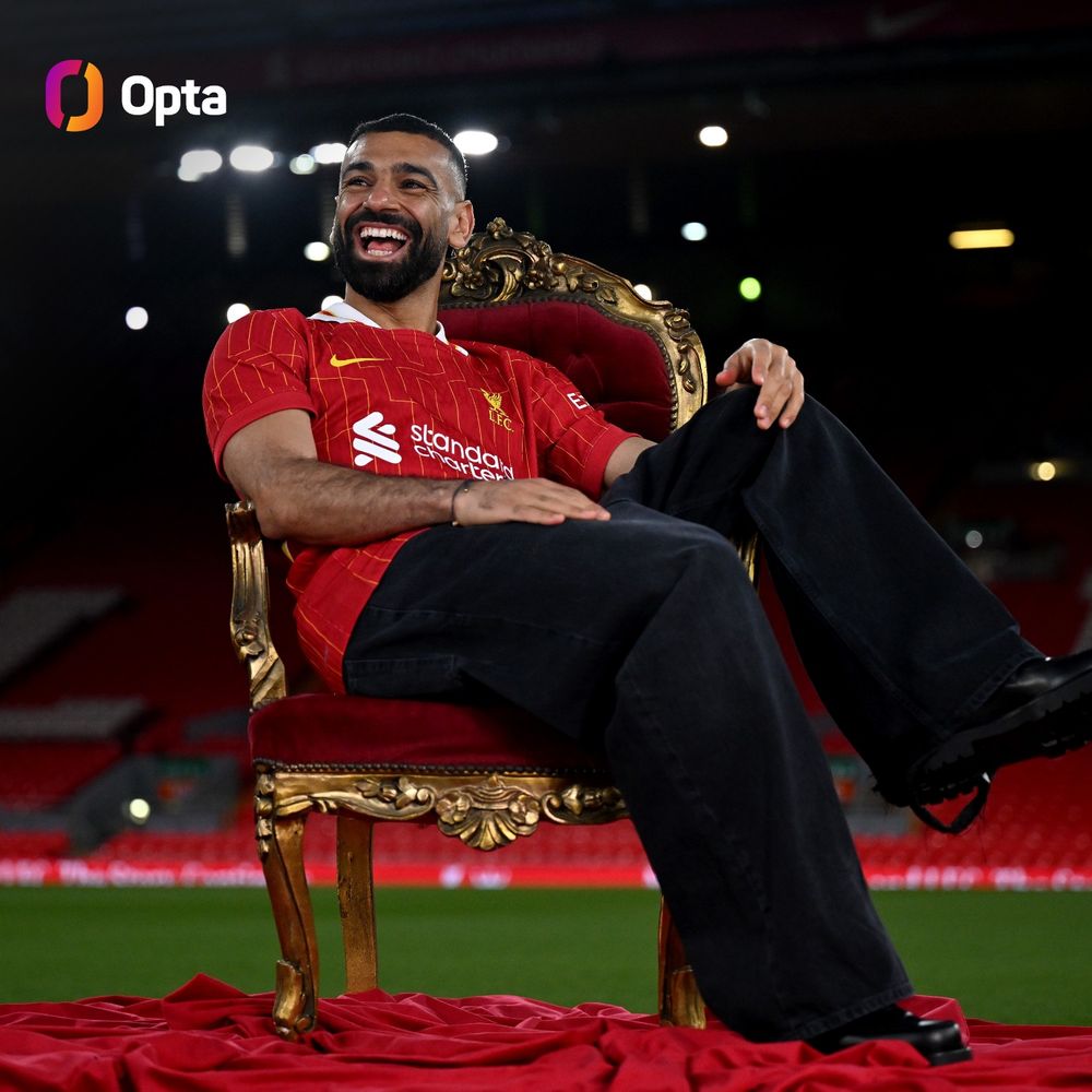 Mo Salah sits on a throne set up in the middle of the Anfield pitch, laughing and smiling during his new contract announcement photoshoot.