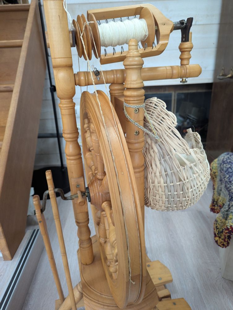 La nouvelle courroie sur mon rouet Kromski Minstrel vu de profil, avec son panier à laine lui aussi fait maison, et une bobine pleine de fil blanc prêt à retordre.