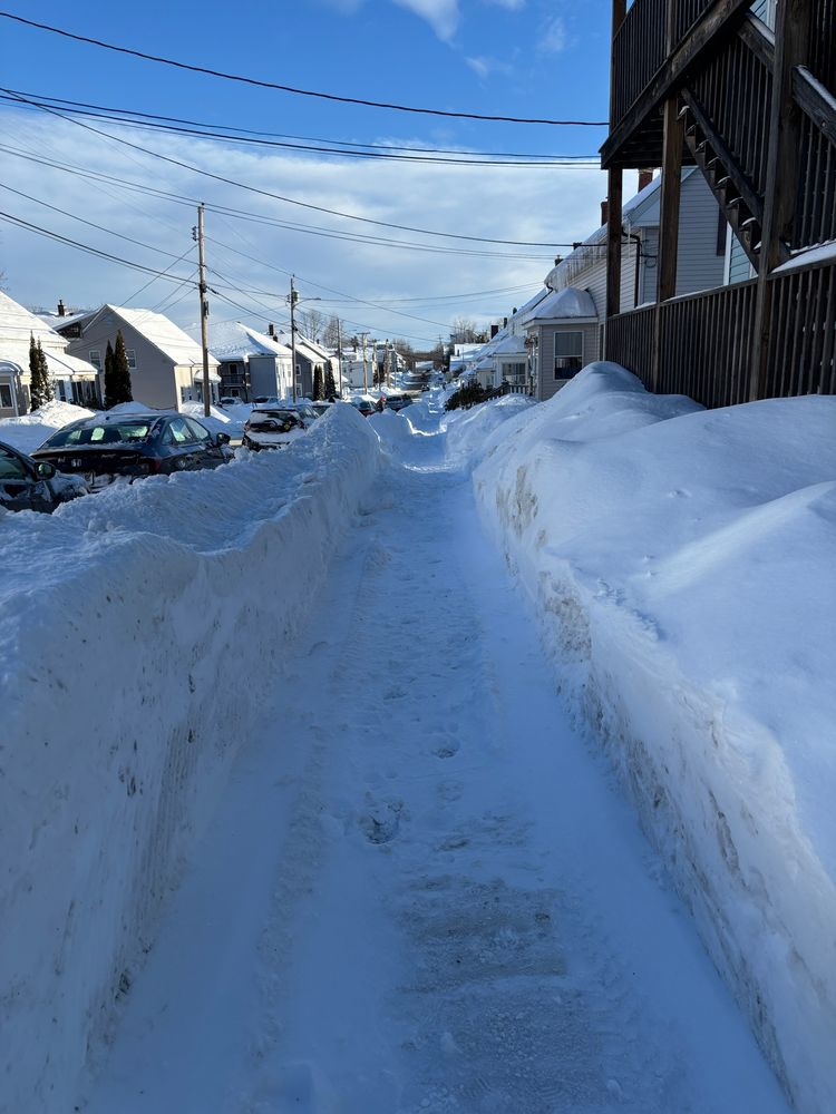 A sidewalk with 2ft walls of snow on either side. Cars on the left past the snow bank. 