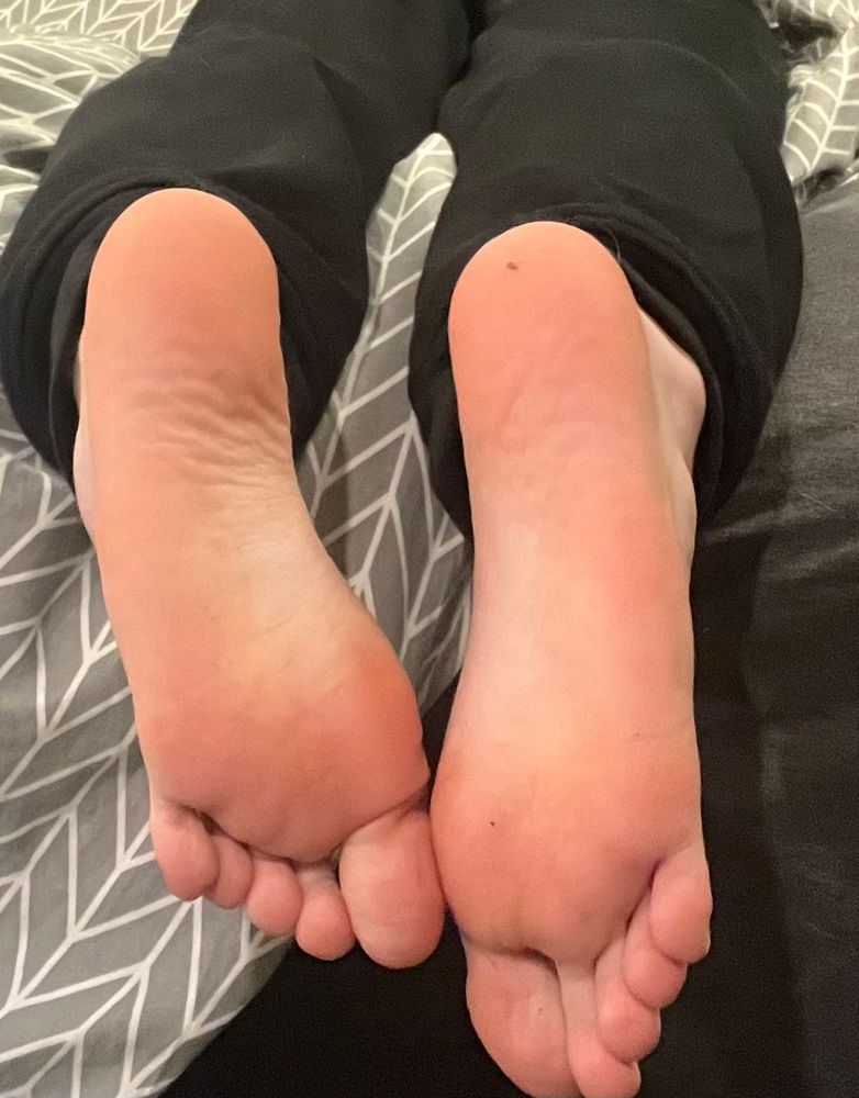 Feet hanging off a bed, soles facing the camera. The sheets are dark grey and the duvet is a light grey and white triangular pattern 