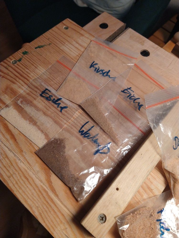Blick auf eine mobile Werkbank, darauf ein Bankhaken zum rechtwinkligen Sägen. Darauf verteilt liegen einige Kunstoffbeitel mit verschiedenfarbigem Sägemehl darin; die Beitel sind von Hand beschriftet: Kirsche, Eiche, Esche, Walnuß.