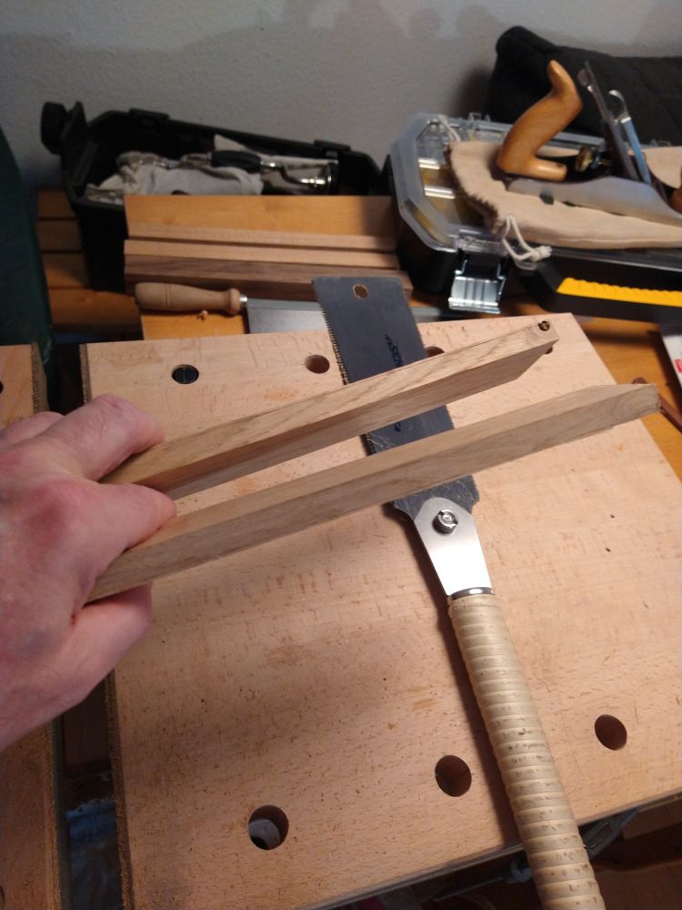 Eine Hand hält zwei gleich dicke Eichenbrettchen in die Bildmitte. Darunter eine mobile Werkbank auf einem Eßtisch, darauf eine Zugsäge nach japanischem Vorbild. Im Hintergrund weitere Holzwerkzeuge.