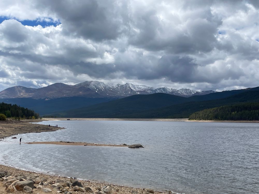 View of an alpine lake in the foreground leading to green hills and snow dusted peaks under clouds in the background