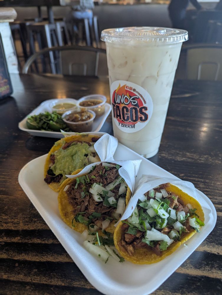 Styrofoam plate with three delicious tacos: El pastor, birria, and pollo.  A GIANT cup of horchata and a treat with three salad and limes sits behind the tacos.  