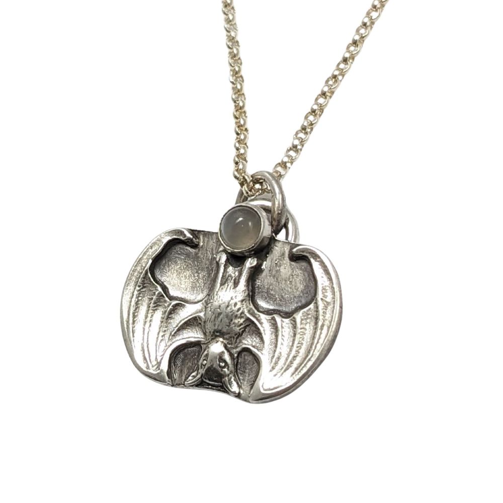 Sterling silver pendant of a bat hanging upside down with it's wings half open. Above the bat there's a gray moonstone gemstone.