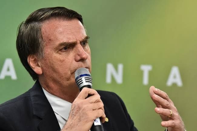 Foto de bolsonaro com microfone na boca e ao fundo ele tapa uma mensagem que faz com que pareça que está escrito atrás dele "anta"