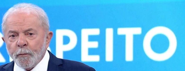 Foto de lula com uma mensagem ao fundo e a cabeça dele tapa de tal forma que faz parecer que está escrito na parede a palavra "peito"