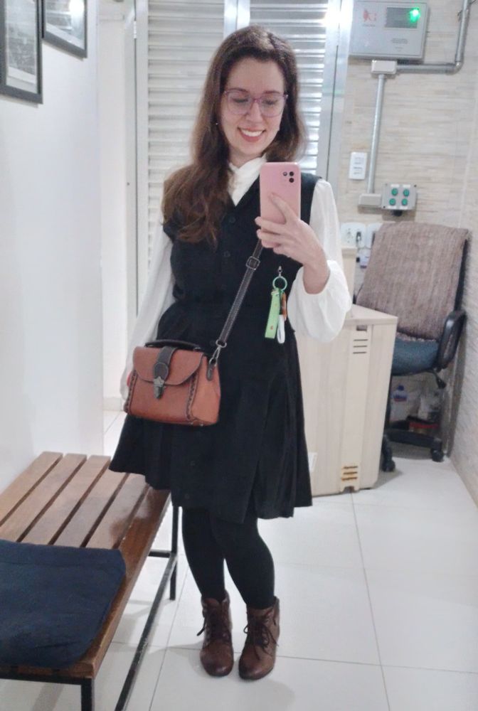 Selfie de corpo inteiro de mulher sorrindo com vestido preto sobre camisa branca meio bufante e meia-calça preta com coturno marrom, com uma bolsa caramelo e marrom à tiracolo.