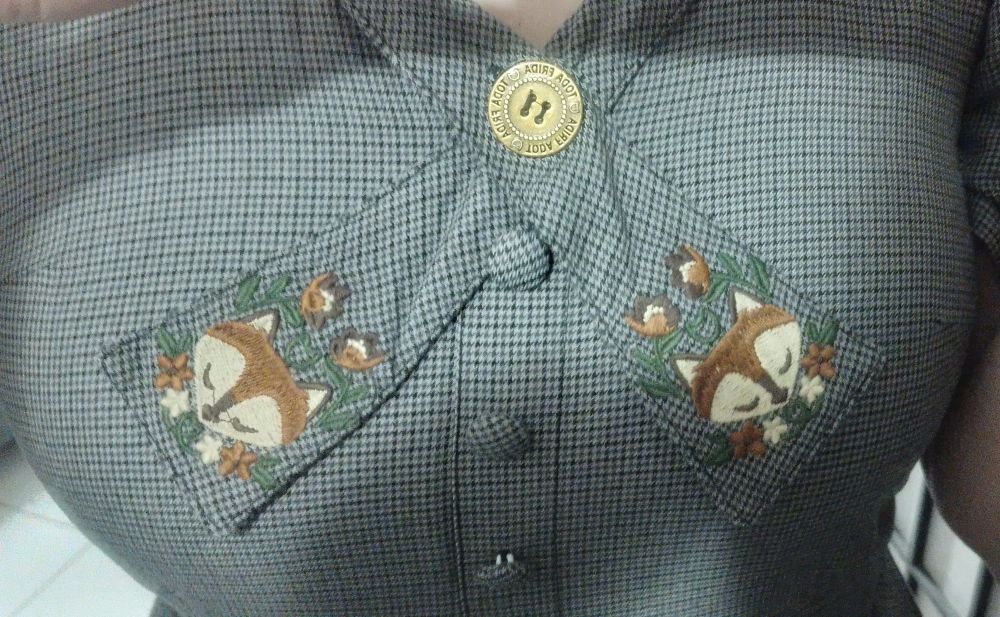 Detalhe do vestido cinza: duas abas/extensões do colarinho ou decote com cabeças estilizadas de raposa bordadas em marrom-alaranjado com flores na mesma cor e folhas verde-escuro.