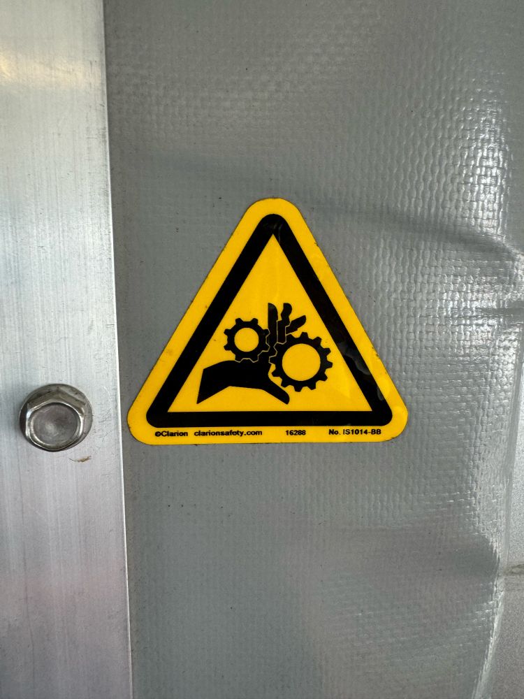 Yellow triangular warning sign showing a hand crunched between gears 