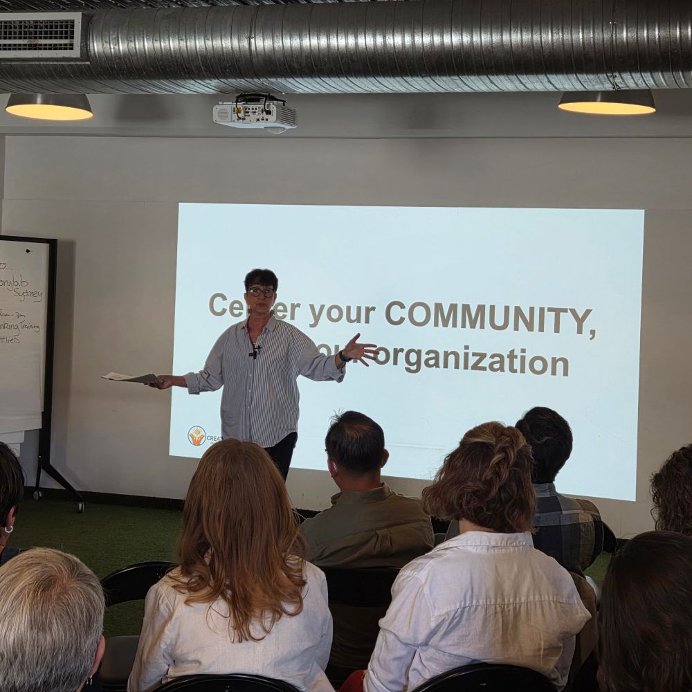 Hildy Gottlieb presenting before a room of people. There's a slide projected on the wall behind her that says "Center your COMMUNITY, not your organization" 