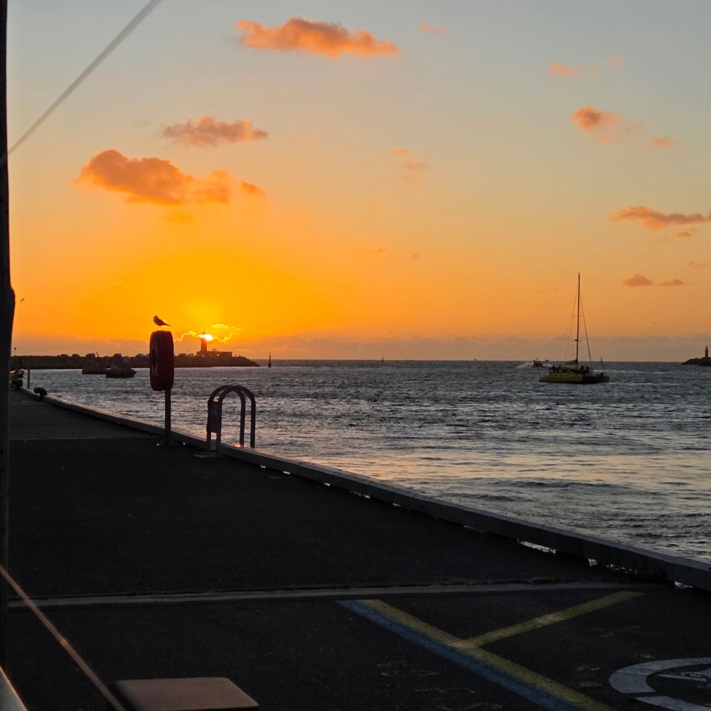 The sun setting into the ocean, immediately behind a small lighthouse at the entry to Fremantle harbour. There's a catamaran in the harbour and a jetty walkway in the foreground. 