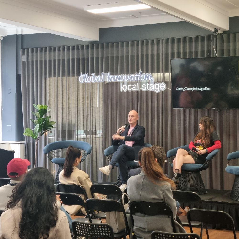 A man and a woman on a small stage in front of rows of folding chairs. The man is talking into a microphone, the woman is listening. 