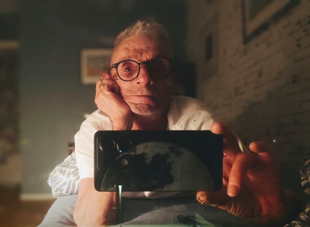 Un homme à lunettes et cheveux blancs courts se tenant la tête sur une main qui fait un selfie allongé sur le ventre sur son lit.